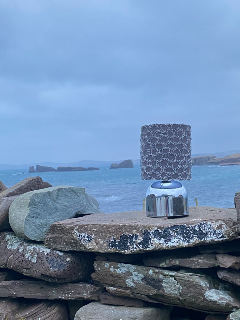a lamp sits on a dry stone dyke with the stacks of Papa Stour and the sea behind it. The lamp has a grey shade that is covered with a knit wire lampshade. 